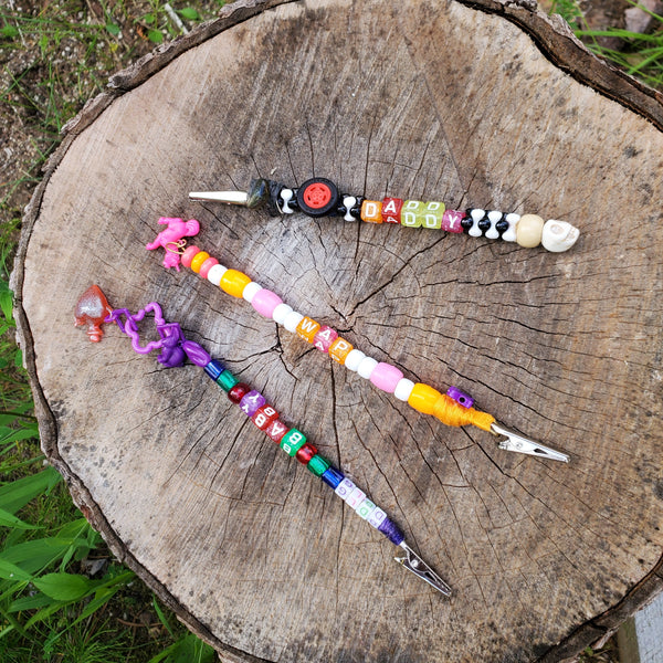 Three colorful beaded clips with wooden and plastic letter beads and small charms are arranged on a cut tree stump outdoors.