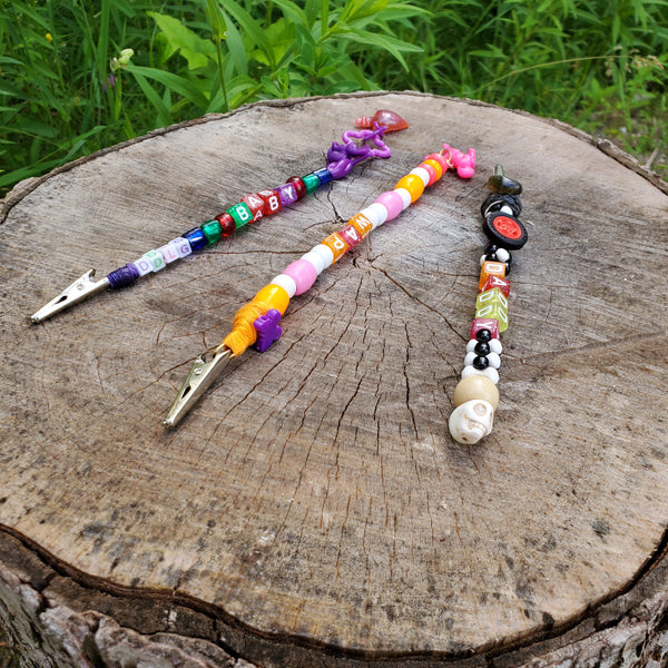 Three colorful beaded metal clips with are arranged on a rustic tree stump outdoors, with green grass visible in the background.