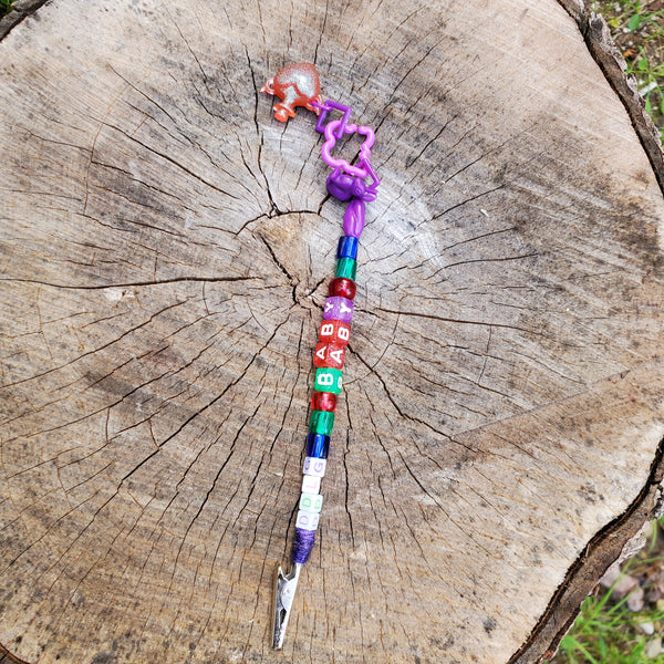A handmade beaded clip, atm clip , bracelet helper tool with a purple plastic ghost, colorful beads, and a small orange heart bottle charm, displayed on a rustic tree stump.