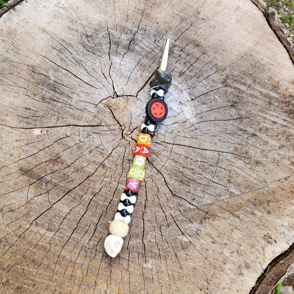 A beaded decorative metal clip or bracelet helper laid diagonally on a weathered tree stump. The metal clip features a white skull bead at the top, black and white patterned beads, a red circular bead, and colorful square beads in yellow, orange, green, pink, and white.