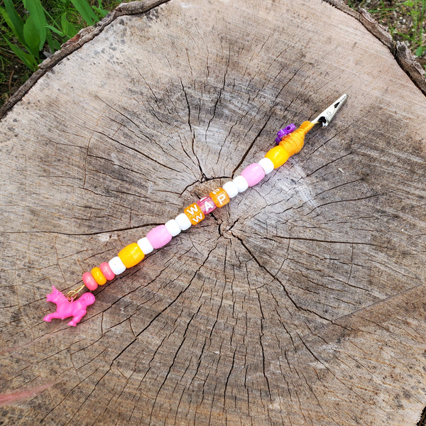 A handmade beaded metal clip displayed on a weathered tree stump. The clip features colorful wooden beads in pink, orange, yellow, and white, with a pink unicorn charm on one end and a purple flower accent near the metal spring clip on the other.