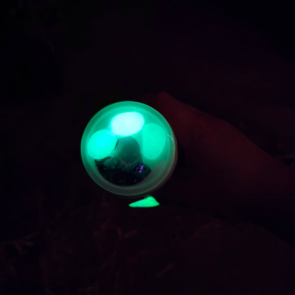 A small round container or cup glowing with bright green bioluminescent-like light, photographed in a very dark environment. A hand is partially visible holding it, and the background is nearly black.