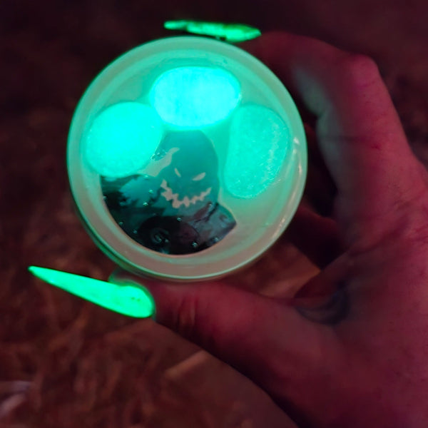 A hand with long neon green fingernails holds a small clear cup or dish containing glowing green substances against a dark brown background. 