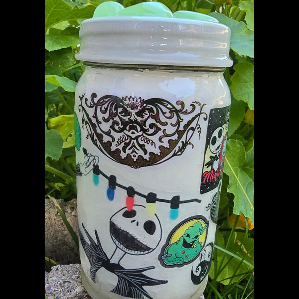 A decorated glass jar with a white lid, featuring hand-painted illustrations of Jack Skellington and other cartoon characters, resting on green leaves and a stone surface.