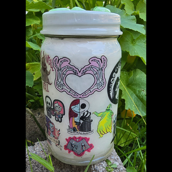 A white ceramic jar with a ribbed screw-top lid, decorated with colorful cartoon-style illustrations, lying on a concrete surface outdoors with green grass and leaves around it.