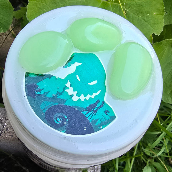 A white plastic jar with a printed sticker of a green monster, filled with three translucent light green gel beads, placed outdoors on a concrete ledge with green leaves and grass in the background.